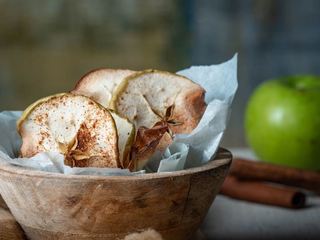 Cinnamon Apple Chips in a wooden bowl with apples and cinnamon sticks in the background. inthekitch.net