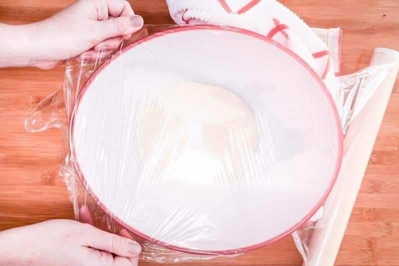 Covering dough in bowl with plastic wrap.