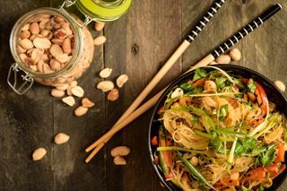 Vegan Pad Thai in a black bowl with chopsticks, peanuts in a jar in the background.