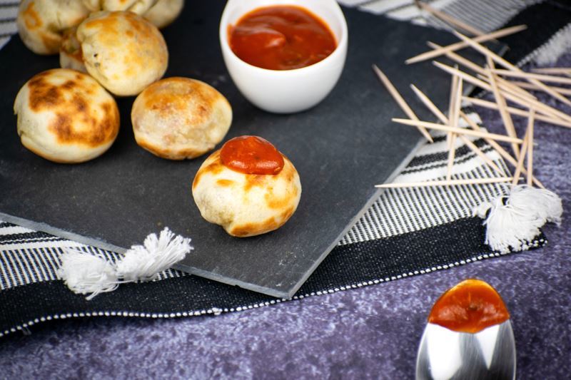 Pizza balls on a black serving plate, small dish of marinara sauce on the side and scattered toothpicks.