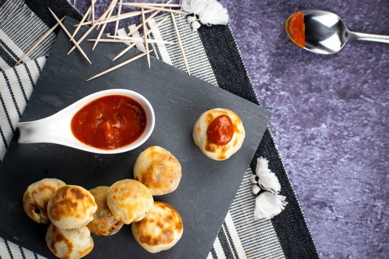 Pizza balls on a black serving plate, small dish of marinara sauce on the side and scattered toothpicks.