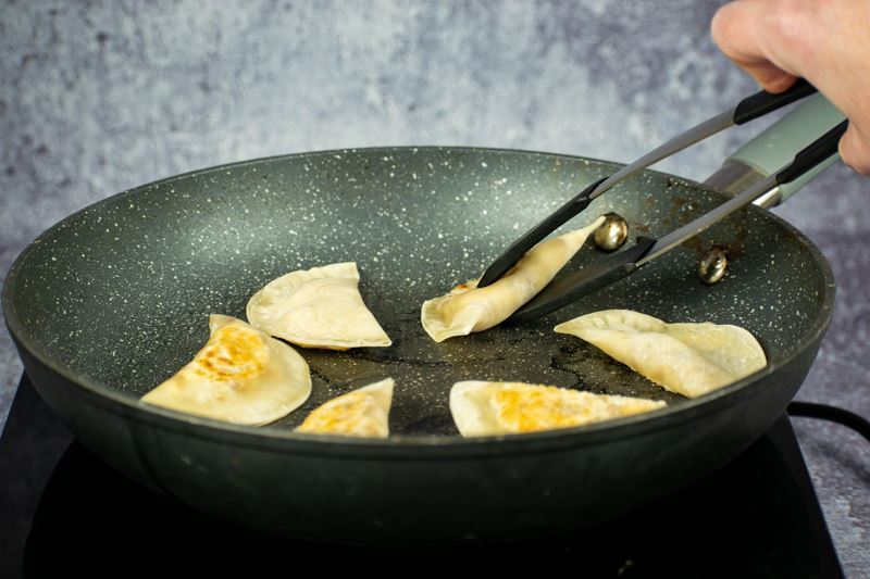 Pierogies cooking in a frying pan, a hand using tongs to flip pierogi.