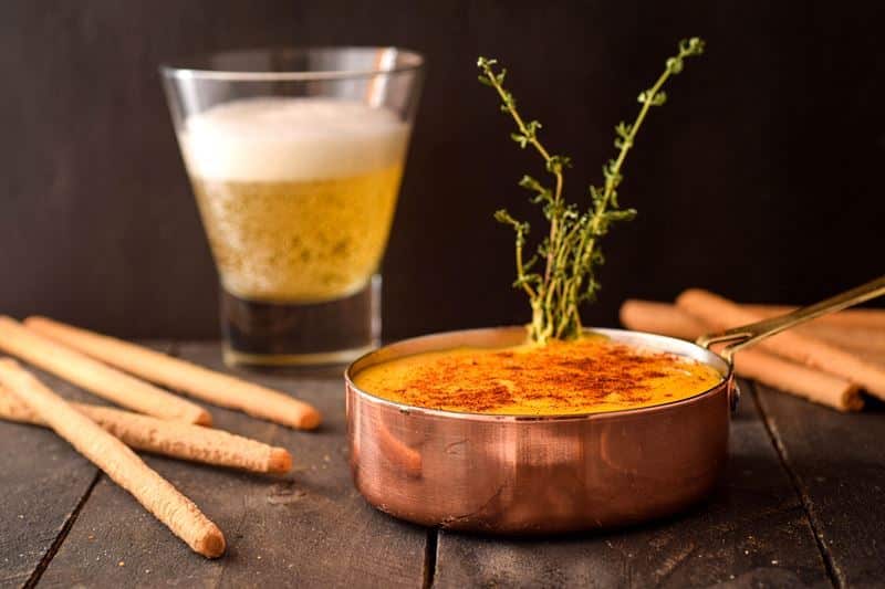 Beer cheese dip in a copper pot with bread sticks and a glass of beer on the side.