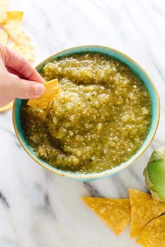 Homemade salsa verde in a bowl, a hand dipping with a tortilla chip.