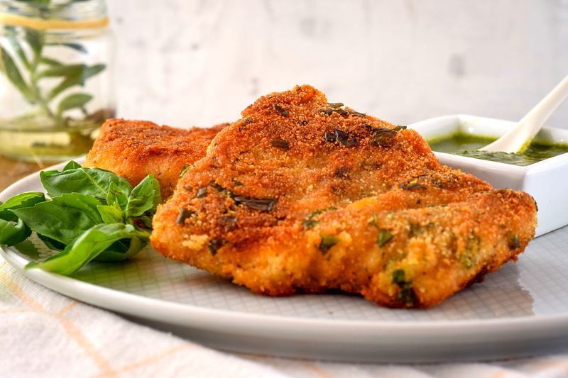 Electric skillet fried fish on a plate with basil pesto in the background and basil leaves on the dish.