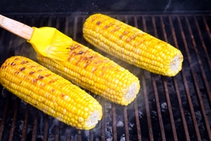 Grilled corn cobs being brushed with butter.