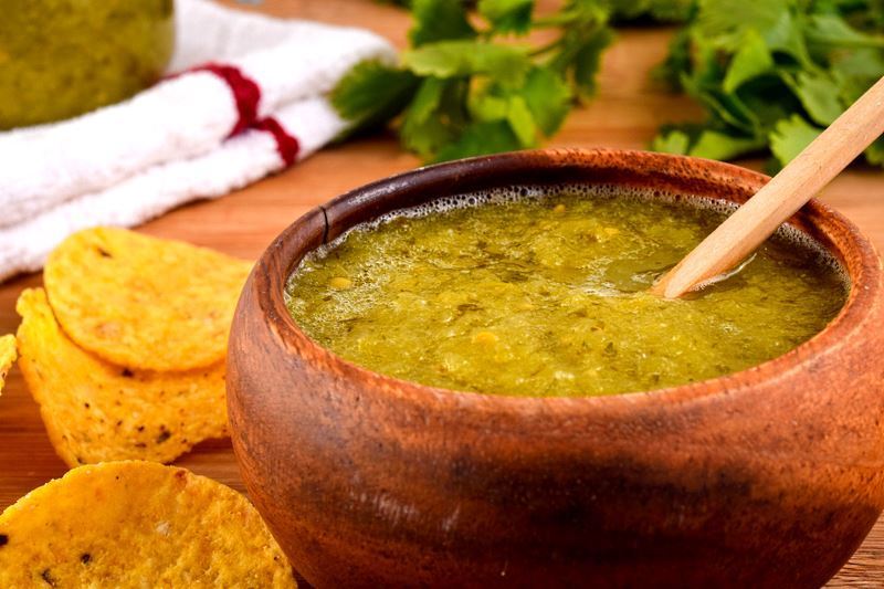 A wooden bowl of tomatillo salsa, a jar of tomatillo salsa, cilantro and tortilla chips in the background.