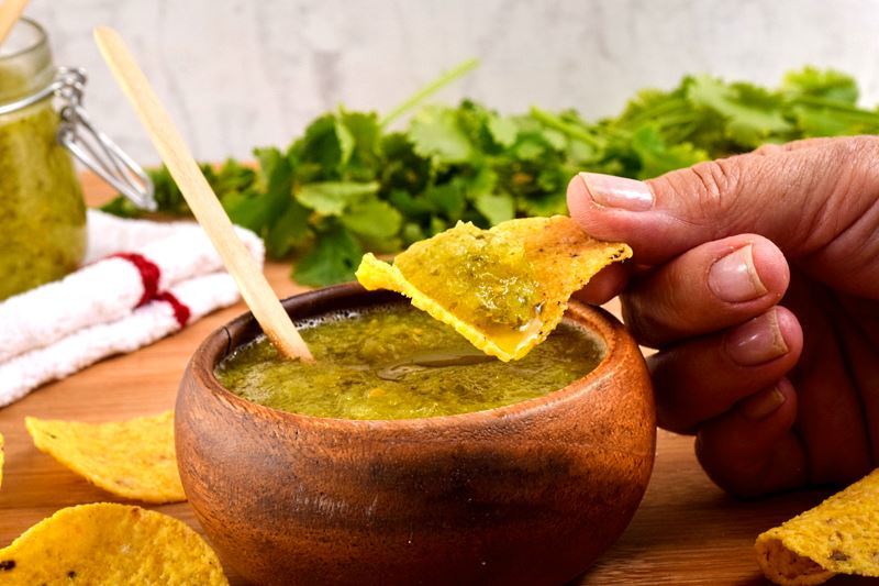 How to Make Tomatillo Salsa_14 A wooden bowl of tomatillo salsa, a jar of tomatillo salsa, cilantro and tortilla chips in the background. Hand dipping a tortilla chip in the salsa.