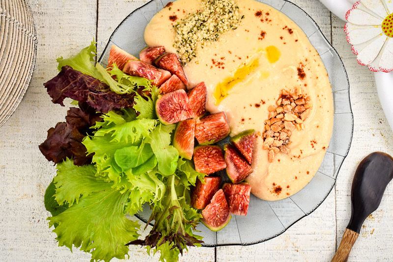 Hummus bowl with fresh figs, greens, nuts, hemp seeds and olive oil on a wooden background. Napkin with a fabric flower and small spatula on the side.