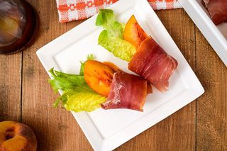 Grilled Peaches Wrapped in Proscuitto on a porcelain appetizer plate, balsamic dressing and peaches in the background.