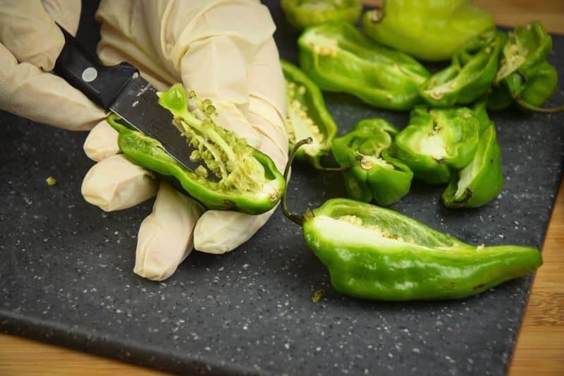 Removing the seeds from a jalapeno pepper sliced in half on a cutting board with a rubber glove.