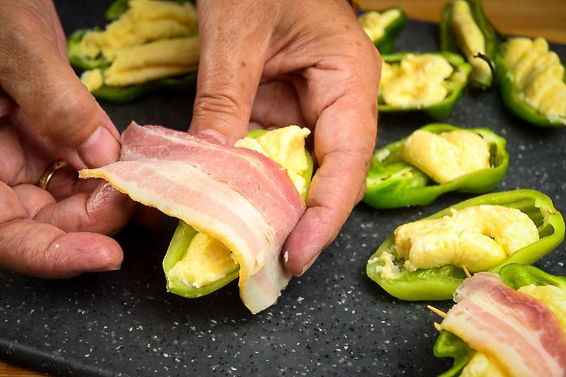A hand wrapping a jalapeno popper with raw bacon on a cutting board.