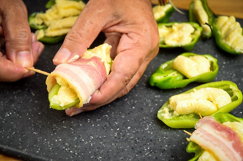 A hand piercing a jalapeno popper with a toothpick, on a cutting board.