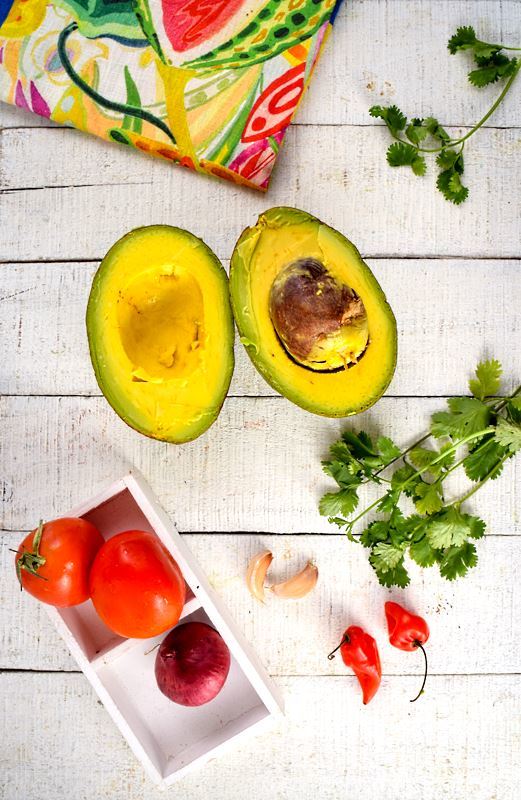 An opened avocado, tomatoes, red onion, serrano peppers, cilantro and a colorful napkin on white wood background.