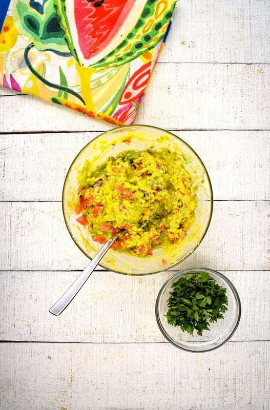 A bowl of guacamole, cilantro and colorful napkin on the side, white wood background.