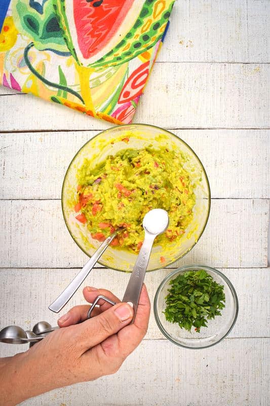 A bowl of guacamole, cilantro and colorful napkin on the side, white wood background. A hand adding salt to the bowl.