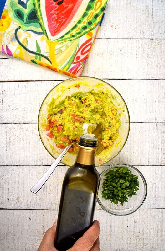 A bowl of guacamole, cilantro and colorful napkin on the side, white wood background. A hand adding olive oil to the bowl.