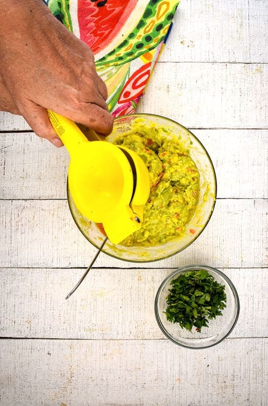 A bowl of guacamole, cilantro and colorful napkin on the side, white wood background. A hand adding lemon juice to the bowl.