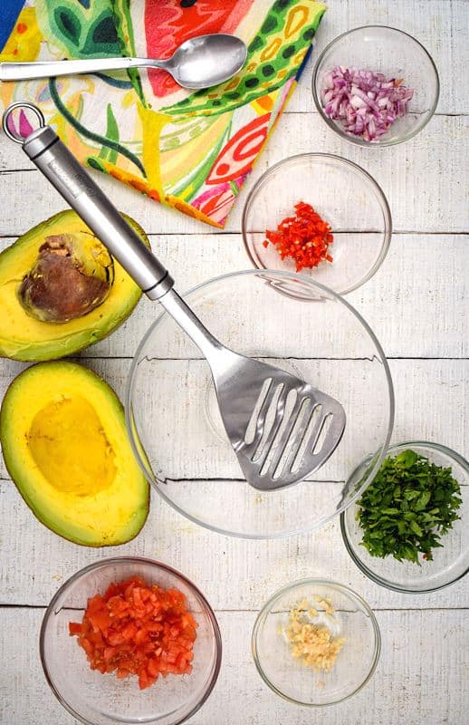 An opened avocado, prepped tomatoes, red onion, serrano peppers, cilantro and a colorful napkin on white wood background.