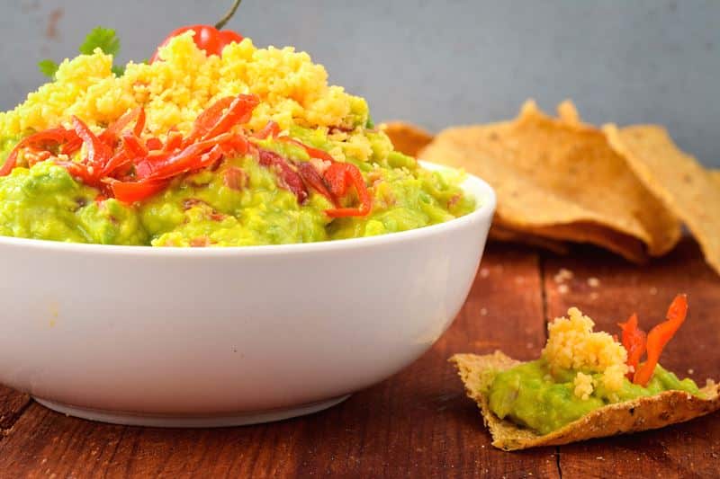 A bowl of guacamole with a chili pepper, cilantro, Mexican seasoning and cheddar cheese. tortilla chips on the side.