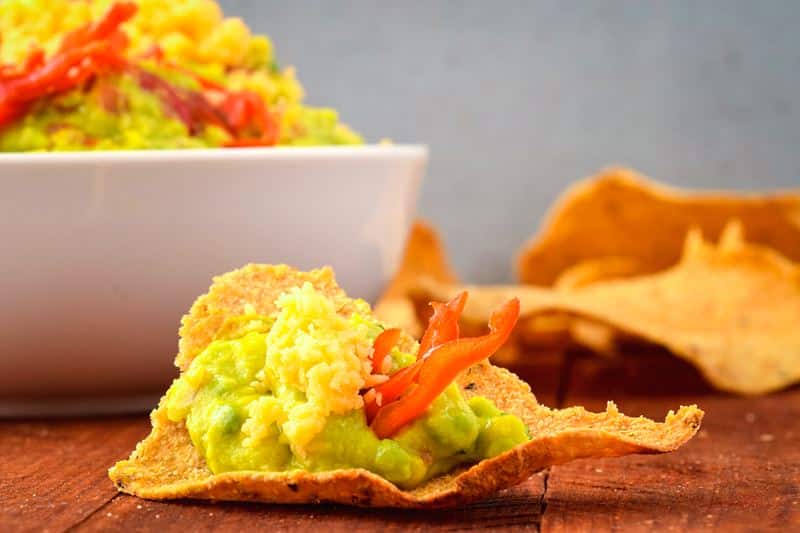 A bowl of guacamole with a chili pepper, cilantro, Mexican seasoning and cheddar cheese. tortilla chips on the side.