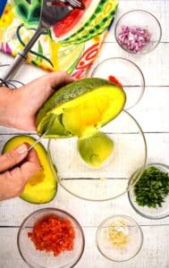 An opened avocado, tomatoes, red onion, serrano peppers, cilantro and a colorful napkin on white wood background. Hands scooping the avocado flesh out.