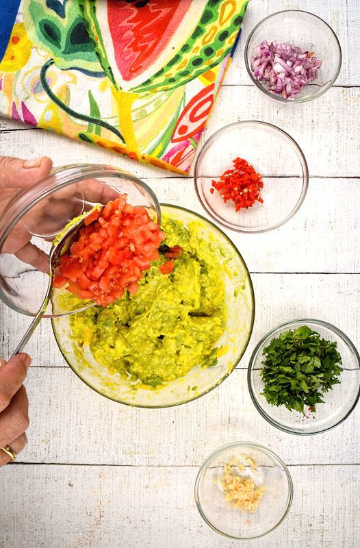 Prepped tomatoes, red onion, serrano peppers, cilantro and a colorful napkin on white wood background. Avocado smashed in a bowl, hand adding the chopped tomatoes.