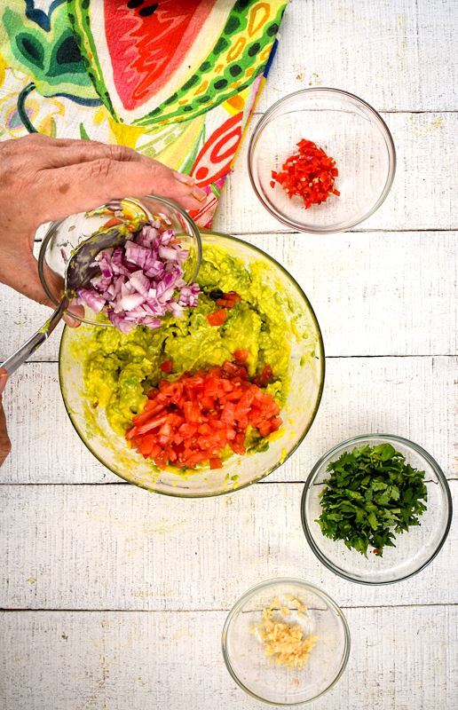 Prepped red onion, serrano peppers, cilantro and a colorful napkin on white wood background. Avocado smashed in a bowl with tomatoes. A hand adding chopped red onion to bowl.