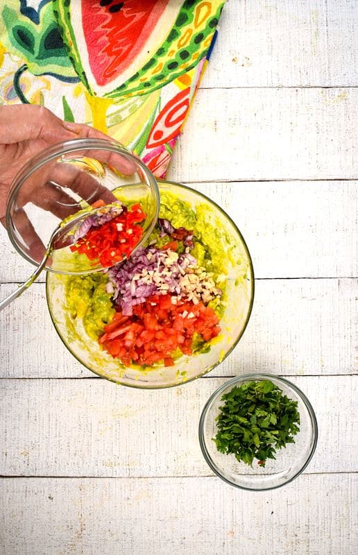 Prepped cilantro and a colorful napkin on white wood background. Avocado smashed in a bowl with chopped tomatoes, red onion and garlic. A hand adding chopped chilies to bowl.