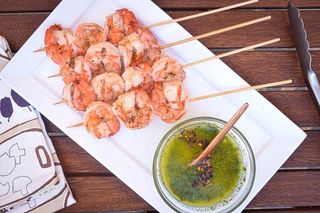 Grilled shrimp skewers on white plate with a bowl of chimichurri sauce.