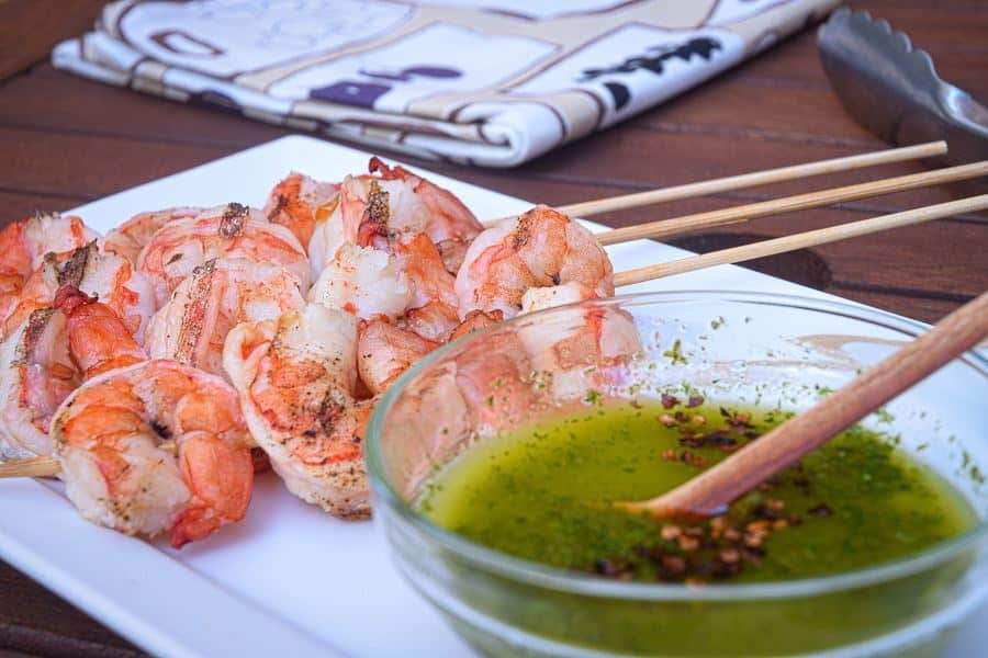 Grilled shrimp skewers on white plate with a bowl of chimichurri sauce.