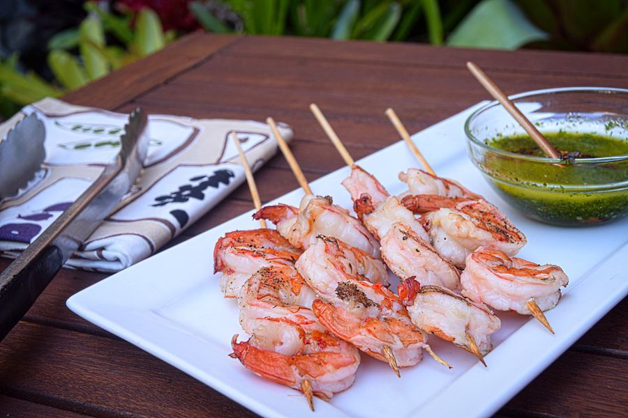 Grilled shrimp skewers on white plate with a bowl of chimichurri sauce.