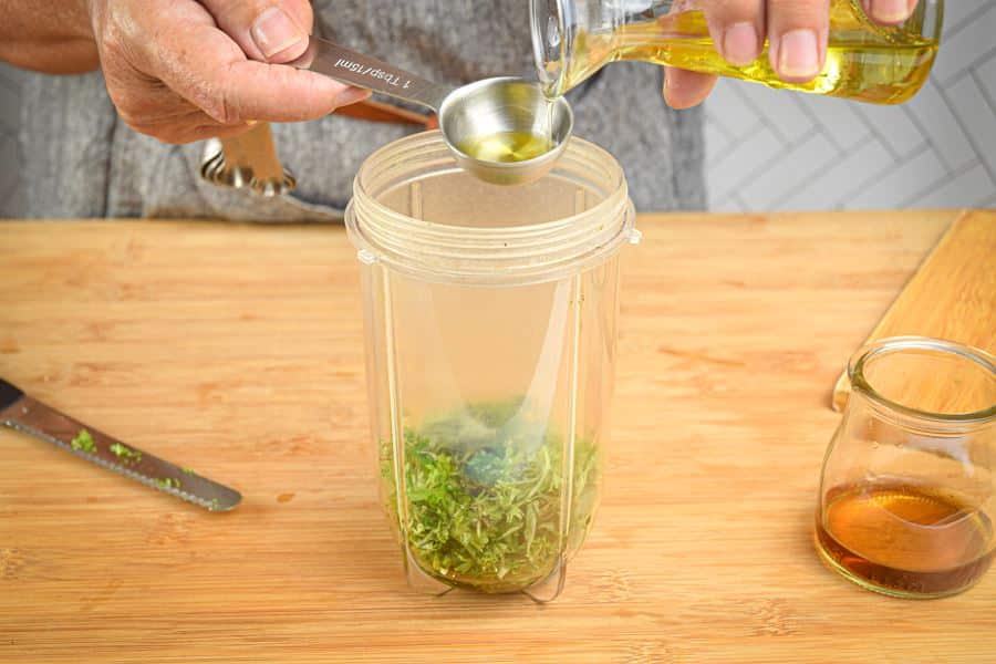A measuring spoon of olive oil pouring into blender jar with parsley.