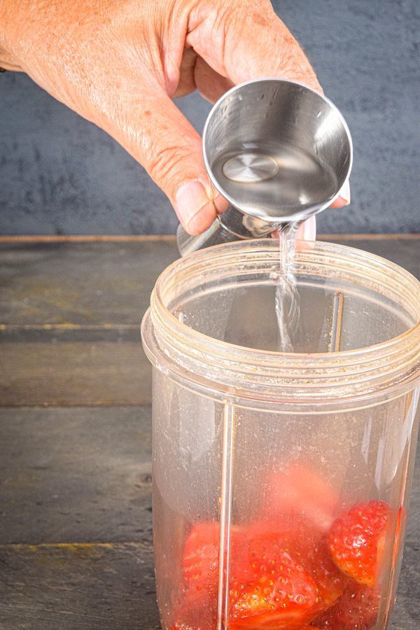 Tequila being poured into blender jar with strawberries and sugar.