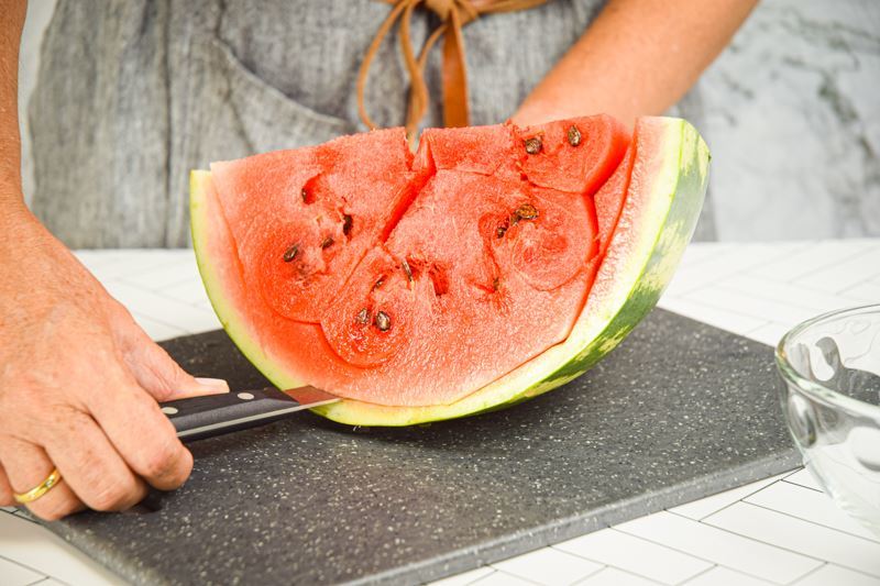 A woman using a knife to peel the rind off the watermelon.
