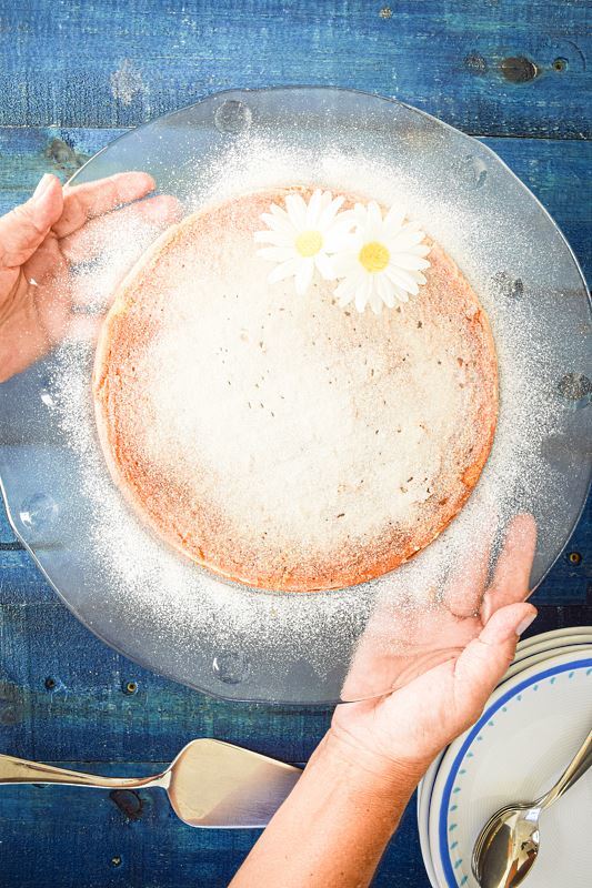 A woman's hand holding a clear plate with electric skillet yogurt cake on it, dusted in icing sugar, 2 white flowers on top, blue wooden background.