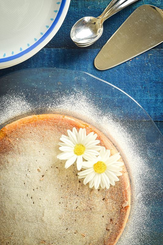 Electric Skillet Yogurt Cake dusted in icing sugar, 2 white flowers on top on a clear plate, blue wooden background, pie server on the side.