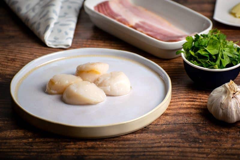 Fresh scallops on a plate, bacon on a plate, garlic and greens in the background.