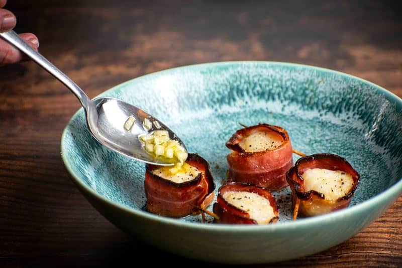 Cooked bacon-wrapped scallops on a blue plate with garlic butter.