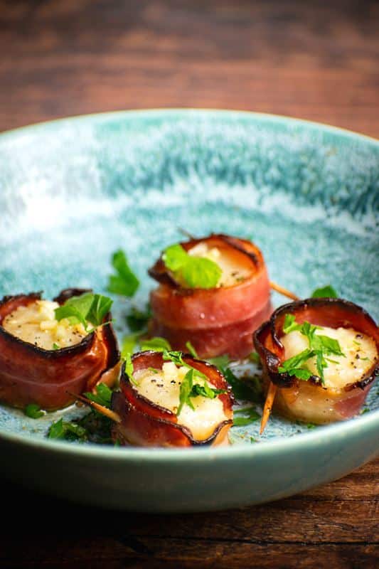 Cooked bacon-wrapped scallops on a blue plate with garlic butter and parsley.