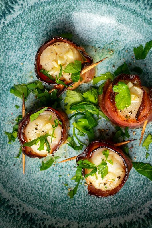 Cooked bacon-wrapped scallops on a blue plate with garlic butter and parsley.