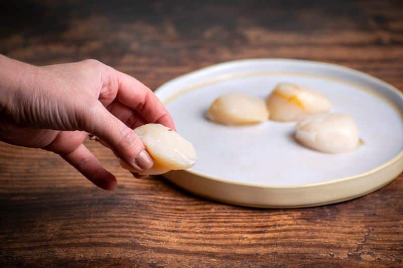 Raw scallops, 1 in a hand, 3 on a plate.