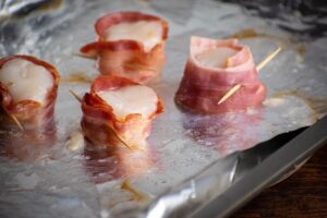 4 scallops on a plate wrapped in bacon and closed with a toothpick, on top of foil-lined pan.