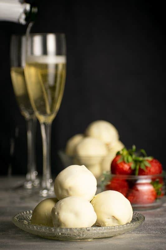 Strawberry Champagne Cake Balls on a clear dish, black background. Glasses of champagne in the background and strawberries.