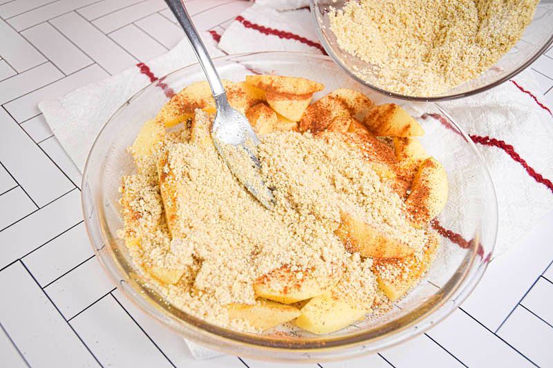 Apple crisp topping ingredients being added to a bowl of apples and cinnamon.
