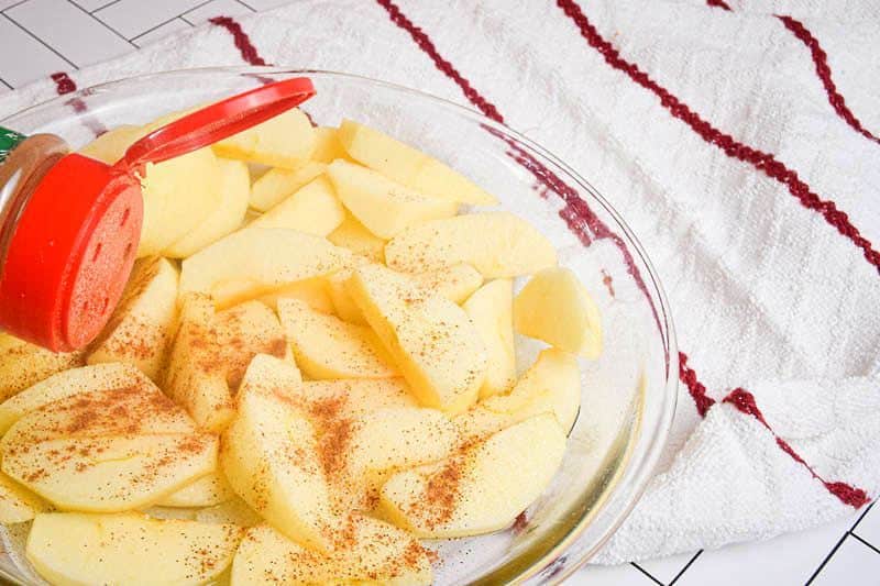 A bowl of sliced applies, cinnamon being sprinkled over them, white towel with red stripes underneath.