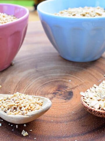 A pink bowl with steel cut oats and a blue bowl of rolled oats with wooden spoons on a wooden background.