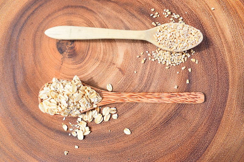 2 spoons filled with rolled oats and steel-cut oats on wooden background.