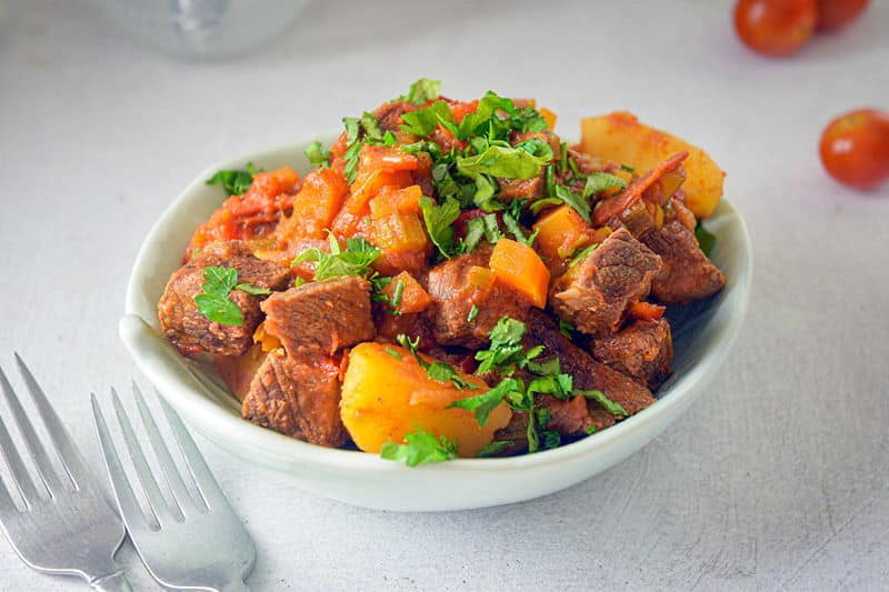 Electric Skillet Beef Stew in a white bowl, cherry tomatoes and two forks on the side, white background.