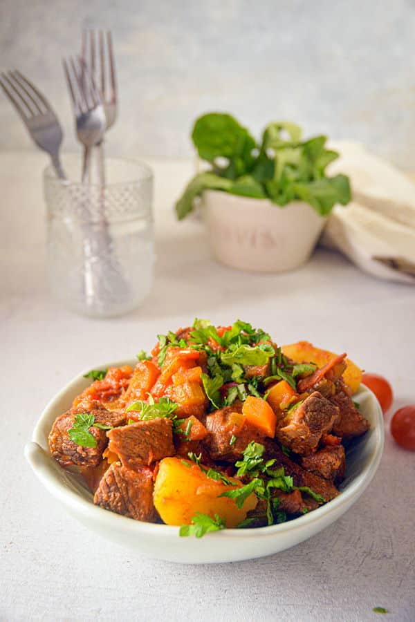 Electric Skillet Beef Stew in a white bowl, cherry tomatoes and forks on the side, white background.